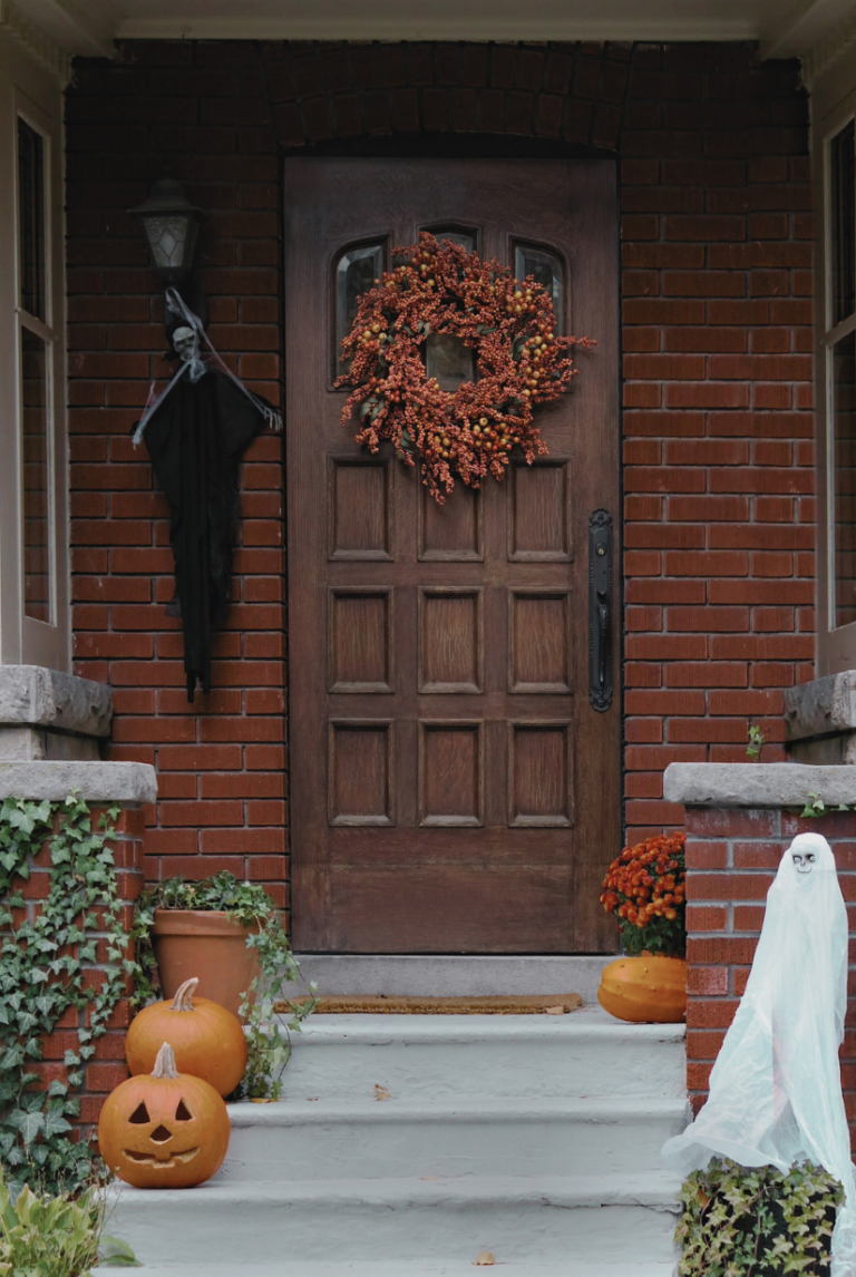 Front porch decorated for Halloween with a rustic wooden door adorned by an orange berry fall wreath, surrounded by carved pumpkins, a hanging skeleton, and a small ghost figure—featured on '30 Affordable Fall Wreath Ideas on Amazon - All Under $50!