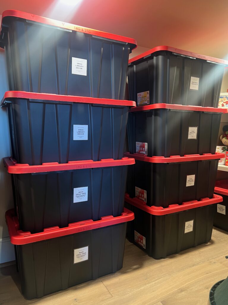 Stacks of large black storage totes with red lids, each labeled on the front with medium-sized white labels, arranged neatly in a storage area.
